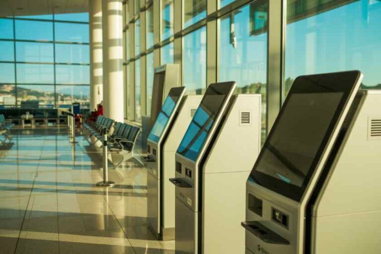 Self-service check-in kiosks in a bright airport terminal with tall windows and seating along the wall