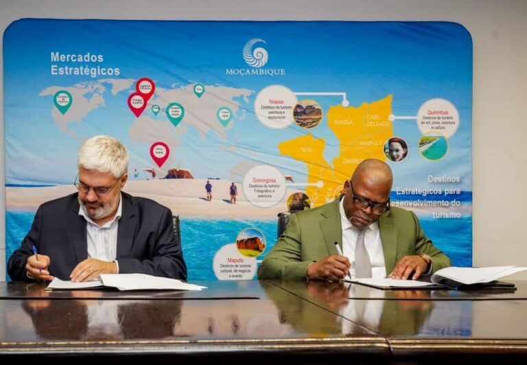 Two men in suits sign documents at a wooden table during a meeting, with a blue Moçambique banner showing a world map in the background behind them.