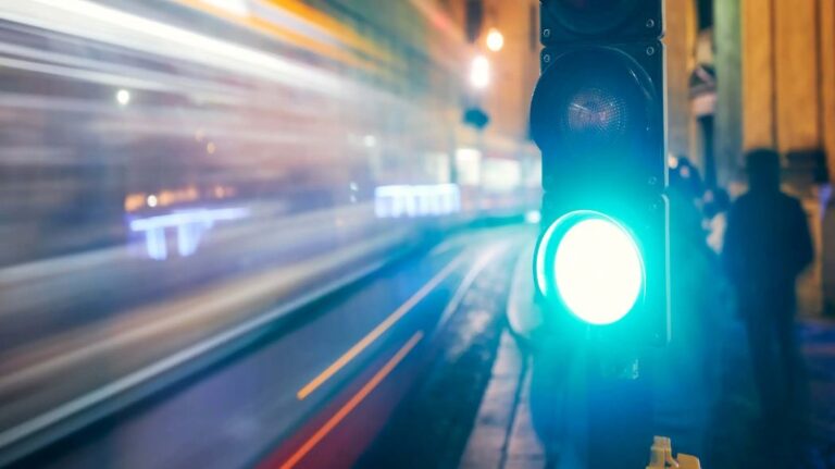 Close-up of a glowing blue-green traffic light at a night city street, with a blurred passing train in the background.