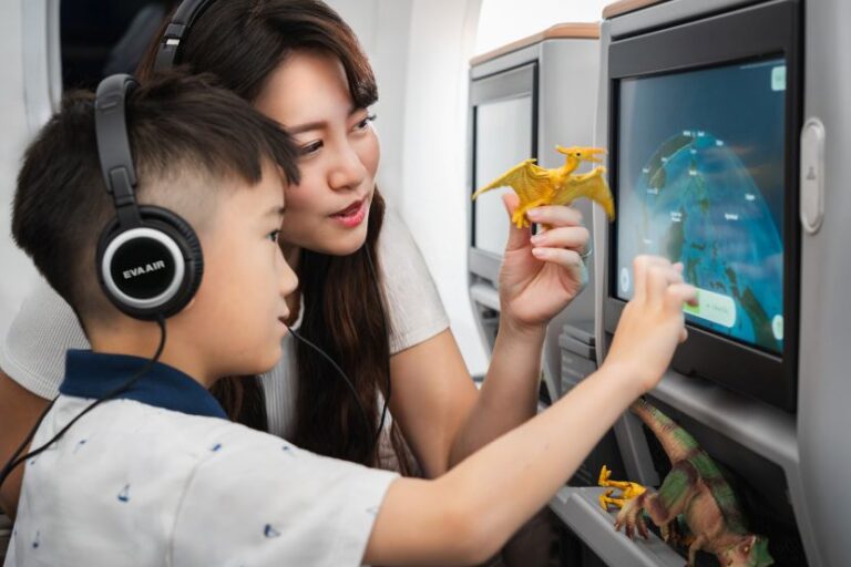 Woman and boy wearing headphones watching an airplane touch-screen and playing with a small yellow dragon toy while seated by the screen.
