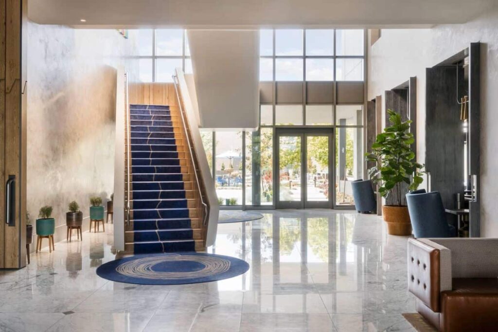 Hotel lobby with a central blue-carpeted staircase, circular blue rug, and marble floor leading to glass doors and outdoor seating area.
