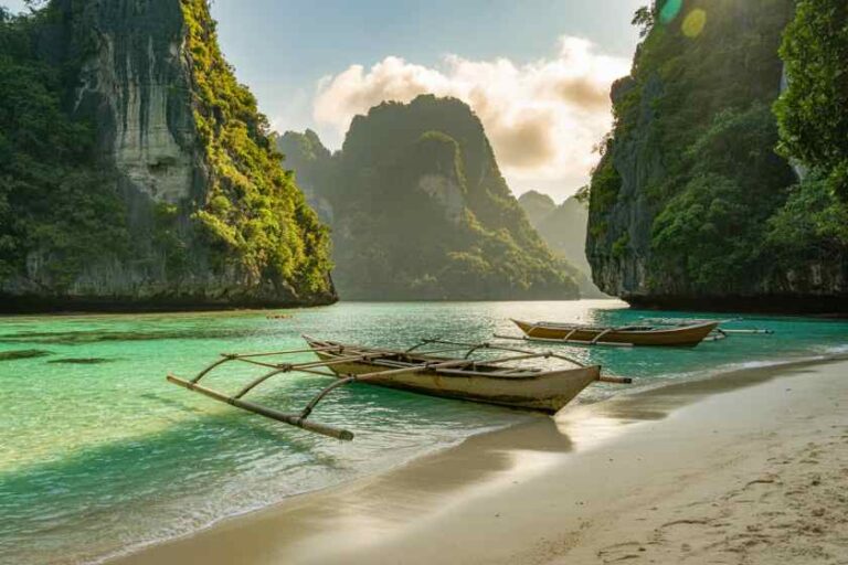 Turquoise sea, white sand, and traditional wooden boats anchored near towering green cliffs. The scene is a calm tropical cove surrounded by limestone formations.