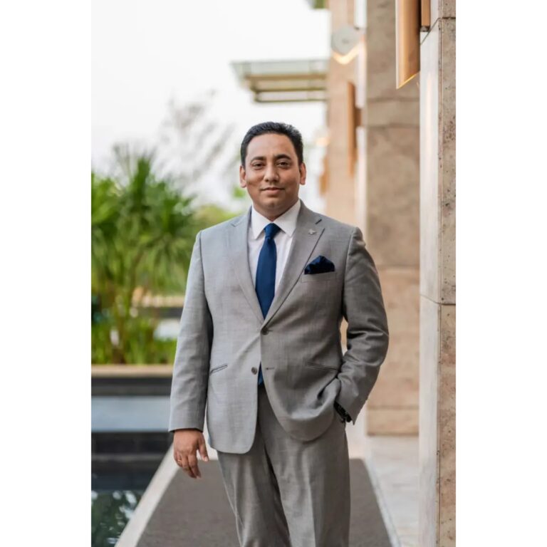 Professional portrait of a man in a gray suit with a blue tie, standing outdoors beside a stone wall.
