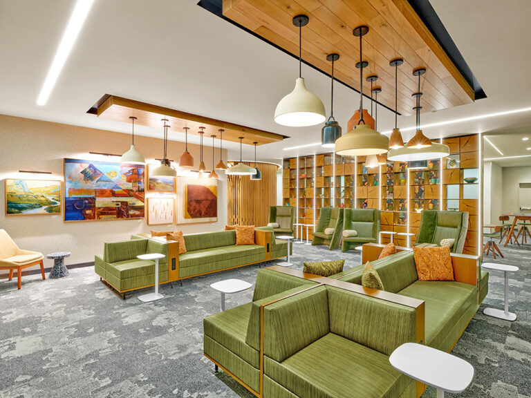 Modern lounge area with green sectional sofas, pendant lights, and colorful wall art on a spacious carpeted floor.