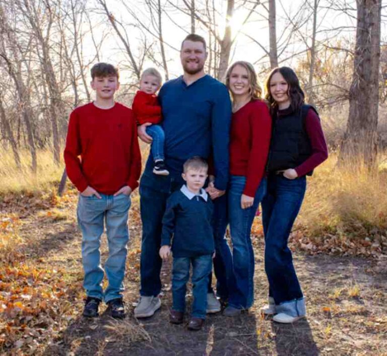 Family of six posing outdoors in autumn woods, smiling at the camera; parents with four children wearing red and blue outfits.