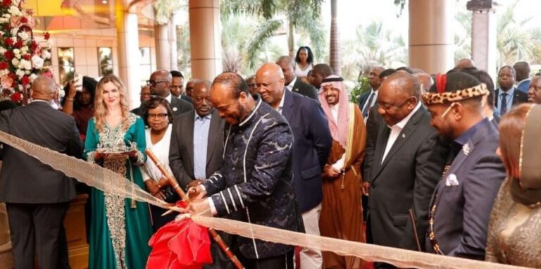 Dignitaries in formal attire cut a ceremonial red ribbon at an official opening amid a crowd of guests and palm trees in the background.