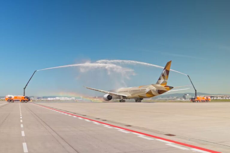 Airliner on a runway receiving a water-spray salute from two orange fire trucks, arching water over the fuselage with a faint rainbow in the background.
