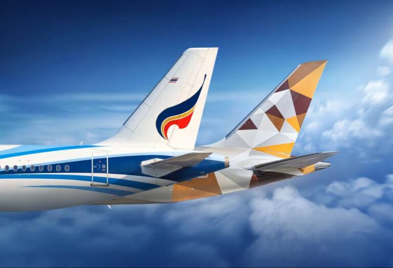Commercial airliner in flight, tail logo visible against a blue sky with white clouds below.