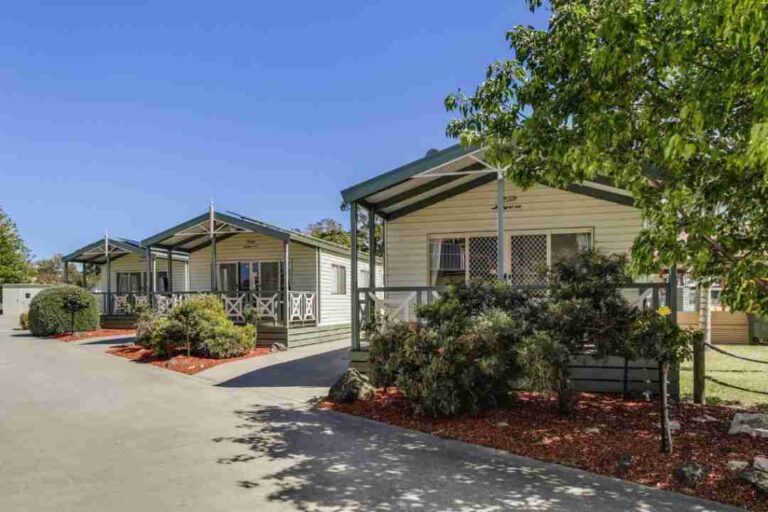 Row of beige mobile homes with covered porches along a paved driveway, landscaped with shrubs and mulch, under a blue sky.