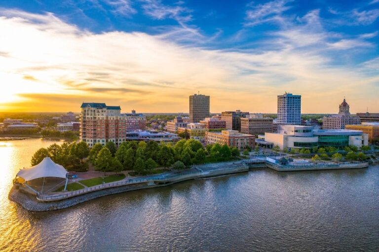 Sunset over a riverfront city skyline with a tree-lined promenade and a white pavilion tent nearby.