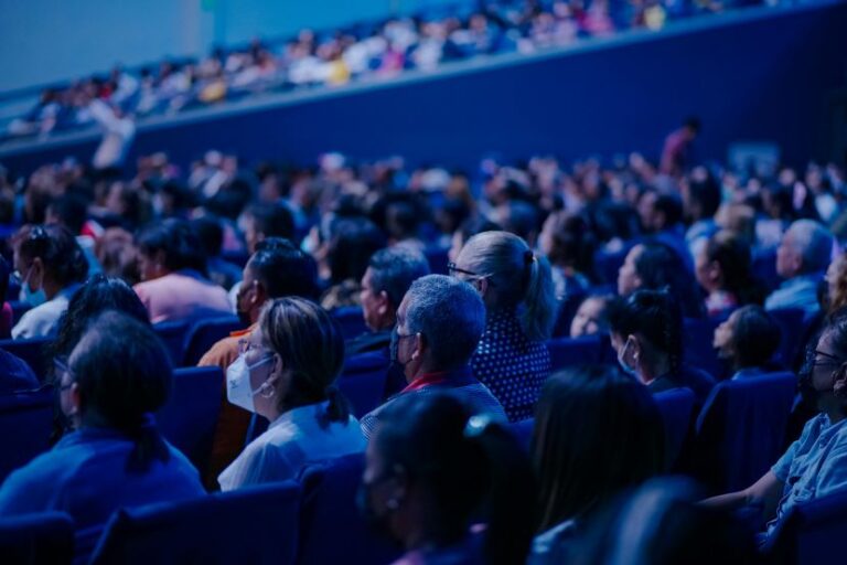 Audience seated in a large, blue-lit auditorium, many wearing masks and watching a performance or presentation.