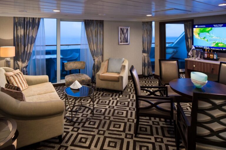 Luxurious cruise ship lounge with beige sofas, armchairs, a round coffee table, and a large wall-mounted TV; balcony doors overlook the sea outside.