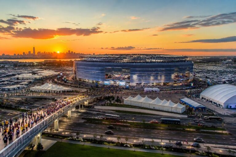 Aerial view of a large stadium at sunset with crowds outside, white tents set up around the venue, and a distant city skyline on the horizon.