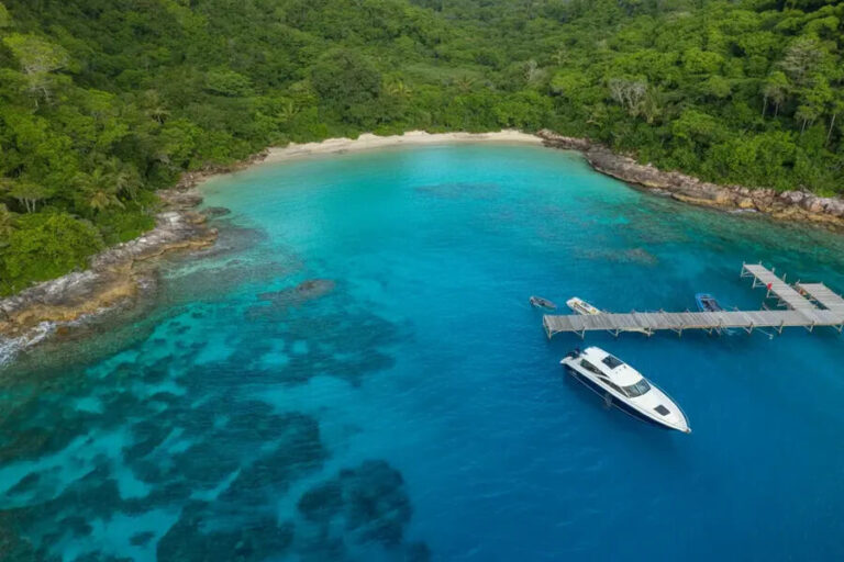 Crystal-clear turquoise cove with a sandy beach and dense green jungle, a wooden pier extending into the water, and a white speedboat moored nearby.