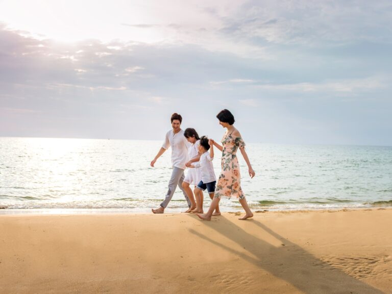 A family of four walks barefoot along a sunny beach, holding hands by the water's edge.