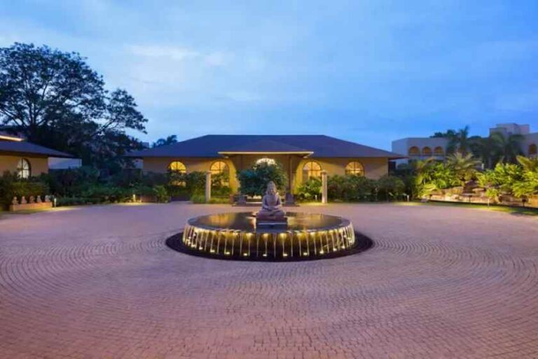 Circular brick courtyard with a lit central fountain, backdrop of arched-window building at dusk.