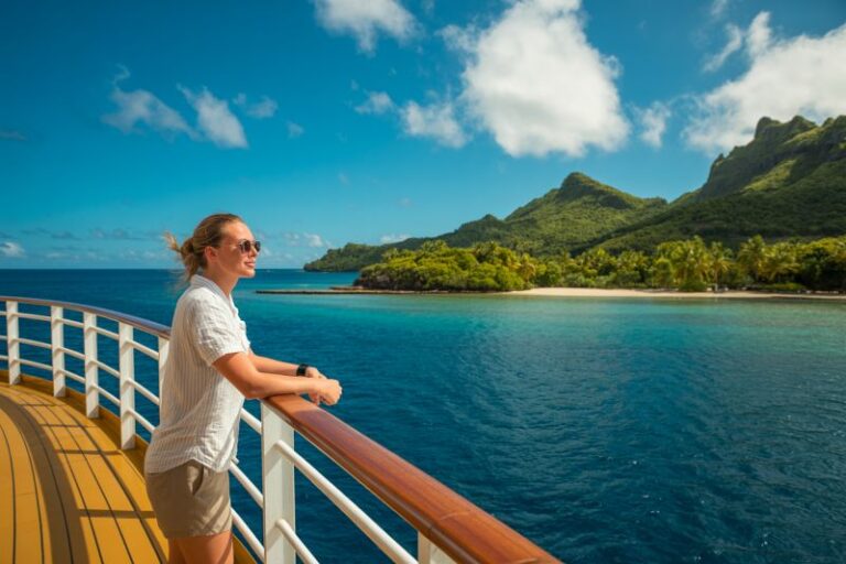 Woman on a cruise deck leaning on a wooden railing, looking at a tropical island with palm trees and blue ocean.