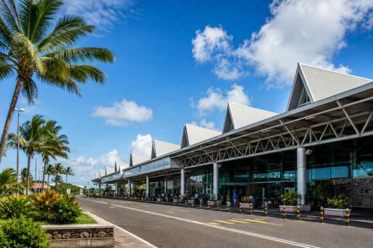 Airport terminal with angular white roofs, glass walls, and palm trees under a blue sky; people near the entrance.