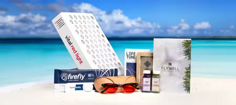 Assorted wellness products and supplements arranged on a sunny beach with turquoise water, including a red-light device, boxes, and sunglasses.