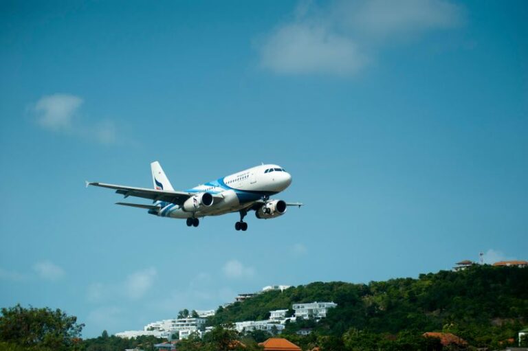 Airplane descending to land over a hillside town with houses and trees under a clear blue sky.