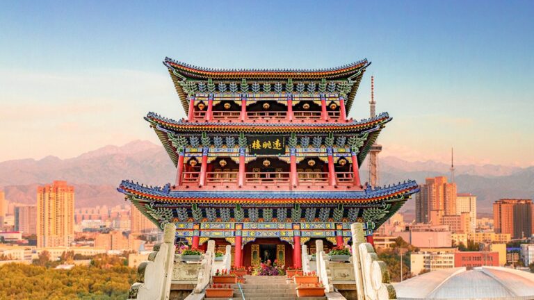 Three-story Chinese pagoda-style temple with ornate red, blue, and gold detailing atop a hill, city skyline and mountains in the background.