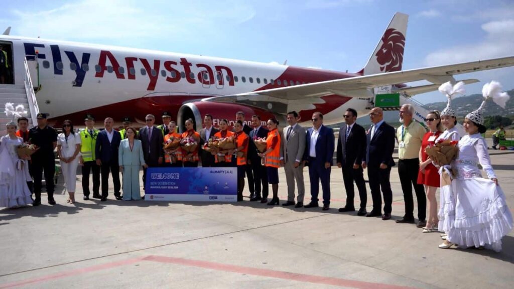 Group of airline staff and officials posing on a tarmac in front of a FlyArystan aircraft during a welcome ceremony, some in orange safety vests and traditional costumes holding flowers and a banner.