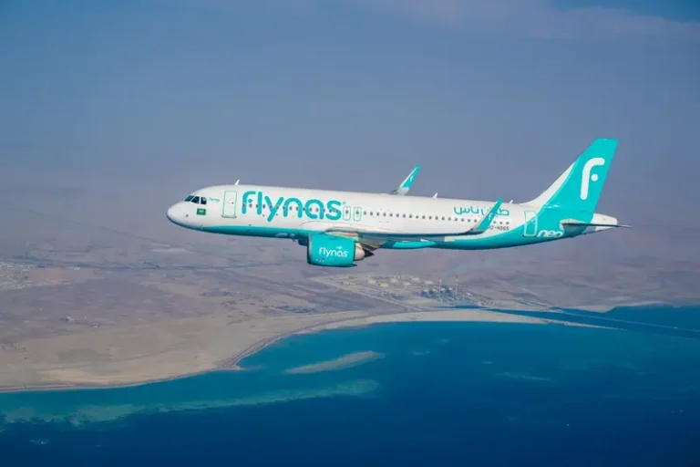 Flynas airplane in turquoise livery flying over a coastline and blue sea.