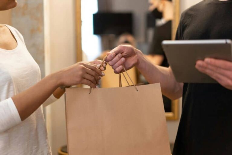 Customer hands a brown shopping bag to a store assistant who is holding a tablet.