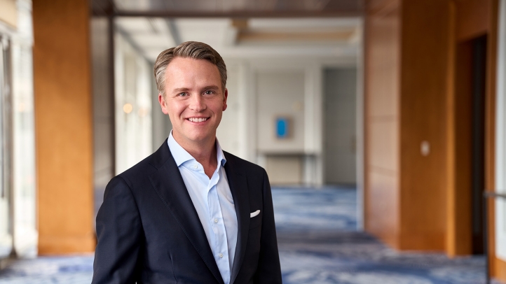 Professional man in a navy suit and light blue shirt smiling in a bright hallway, posing for a business headshot)