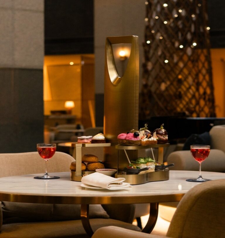 Round marble table with a gold tiered dessert stand holding pastries and sandwiches, two red drinks, and a napkin in a cozy lounge.
