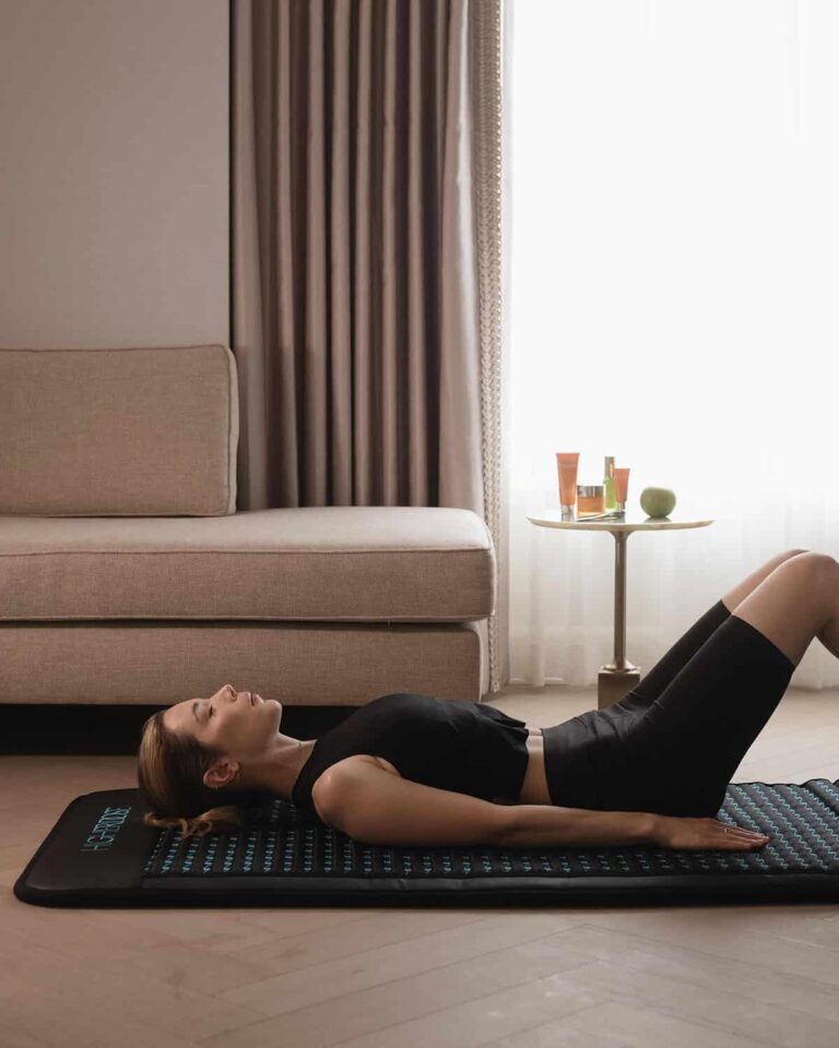 Woman in black athletic wear lying on her back on a yoga mat, performing a glute bridge in a living room near a sofa and side table.