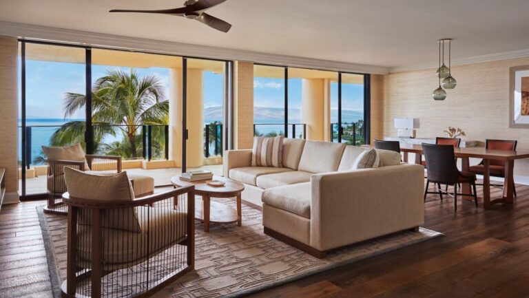 Bright coastal living room with beige sofa and chairs, glass doors opening to a balcony overlooking the ocean and palm trees.