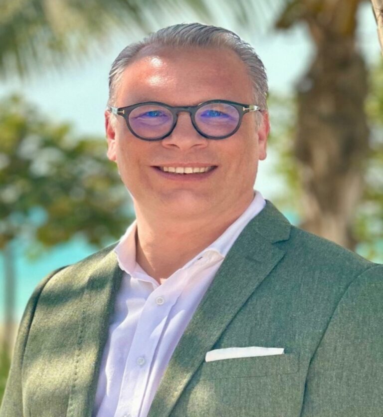 Smiling man in a green blazer and white shirt outdoors with palm trees in the background, wearing round glasses and a white pocket square.