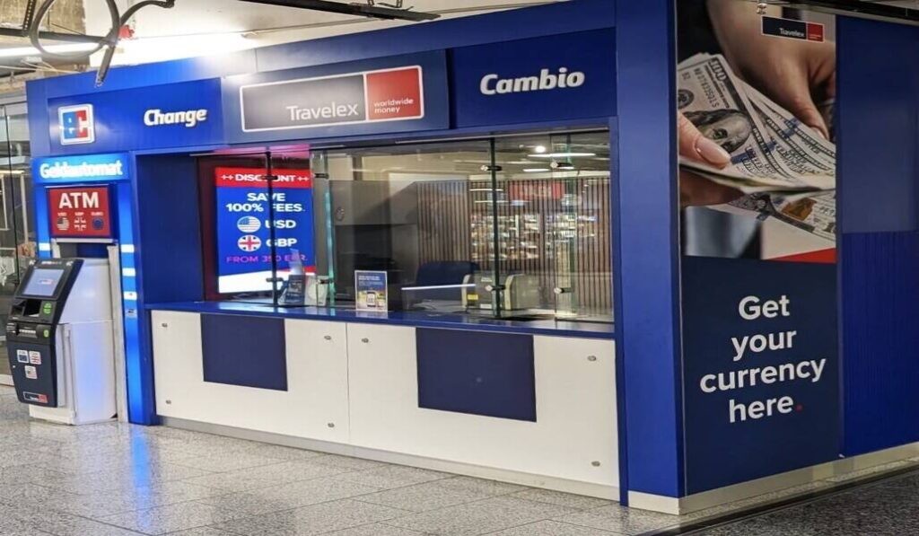Blue currency exchange booth in an airport, with Change/Travelex signs and a large 'Get your currency here' panel.