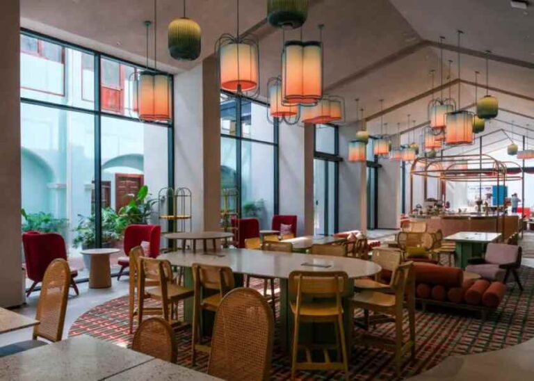 Open, modern cafe interior with large windows, colorful hanging lamps, and mixed seating around round tables.