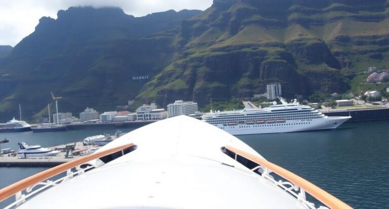 Forward view from a cruise ship bow over calm harbor, with a large white cruise ship and green mountains in the background.