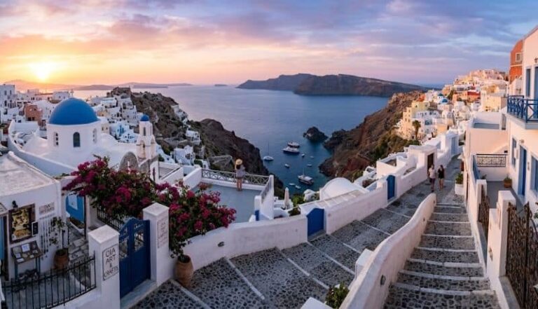 Sunset over white-washed buildings with blue domes in Santorini, overlooking the caldera and boats.