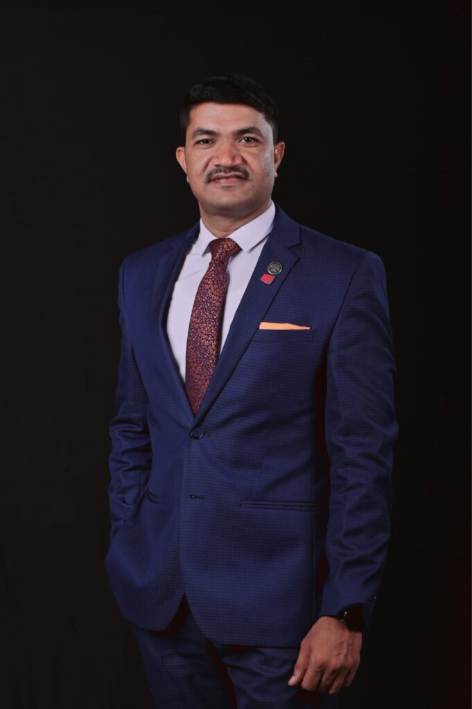 Professional portrait of a man in a navy suit, white shirt, and orange patterned tie, standing against a dark background with a matching pocket square and pin on the lapel.