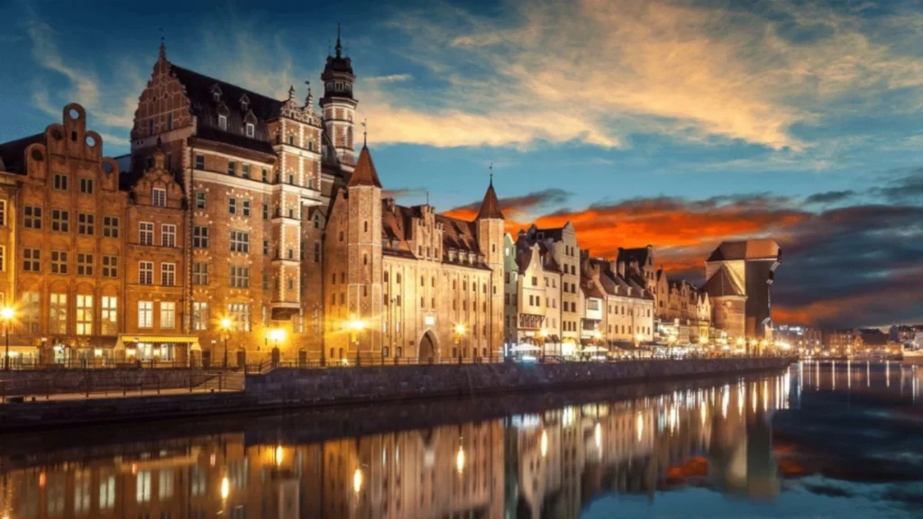 Nighttime view of a row of medieval brick buildings along a river, warm streetlights reflecting on the water.