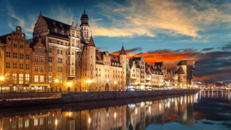 Nighttime view of a row of medieval brick buildings along a river, warm streetlights reflecting on the water.