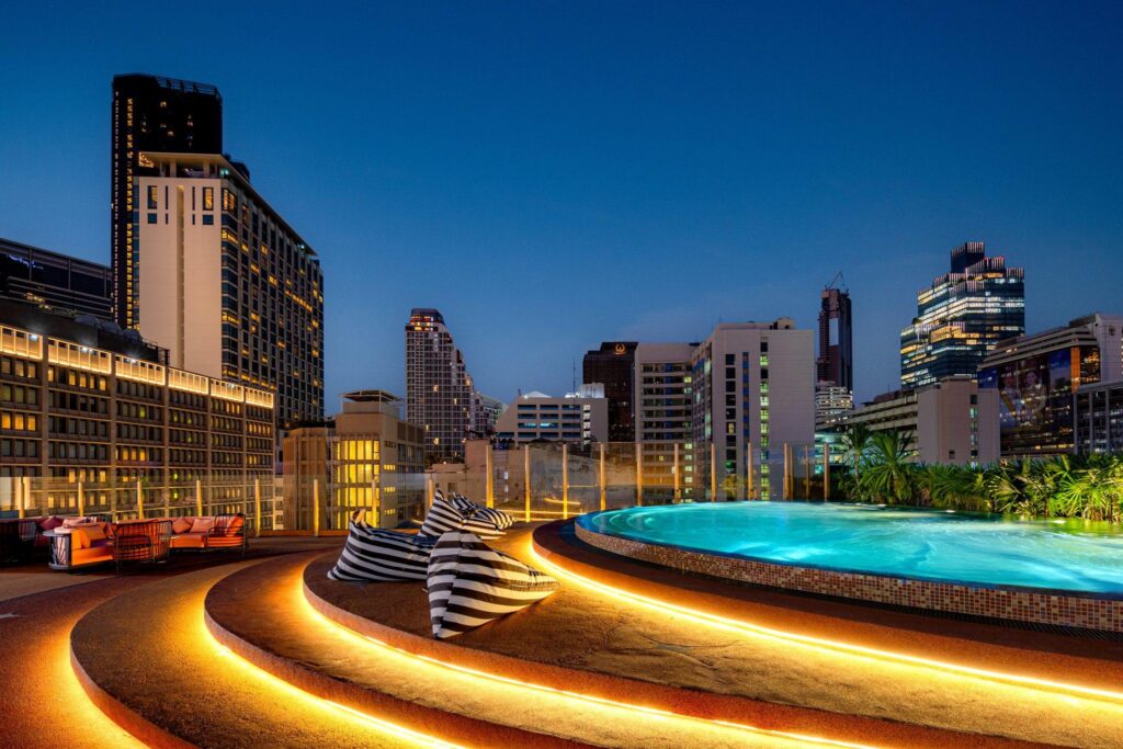 Rooftop pool at dusk with curved illuminated steps, striped lounge cushions, and a city skyline in the background.