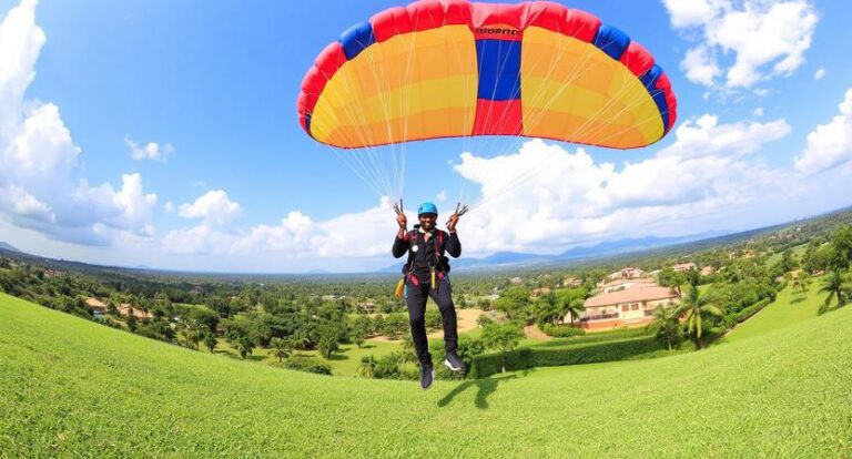 Person wearing helmet and harness paragliding over a green hillside with houses in the distance under a blue sky.