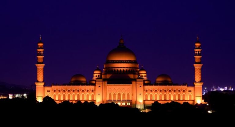 Nighttime view of a grand mosque with a large central dome and two minarets, illuminated in orange against a dark blue sky, with trees in the foreground and city lights in the distance.