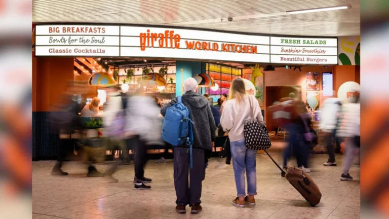 Busy indoor food court with a bright 'giraffe world kitchen' sign; travelers push luggage and stand in line.