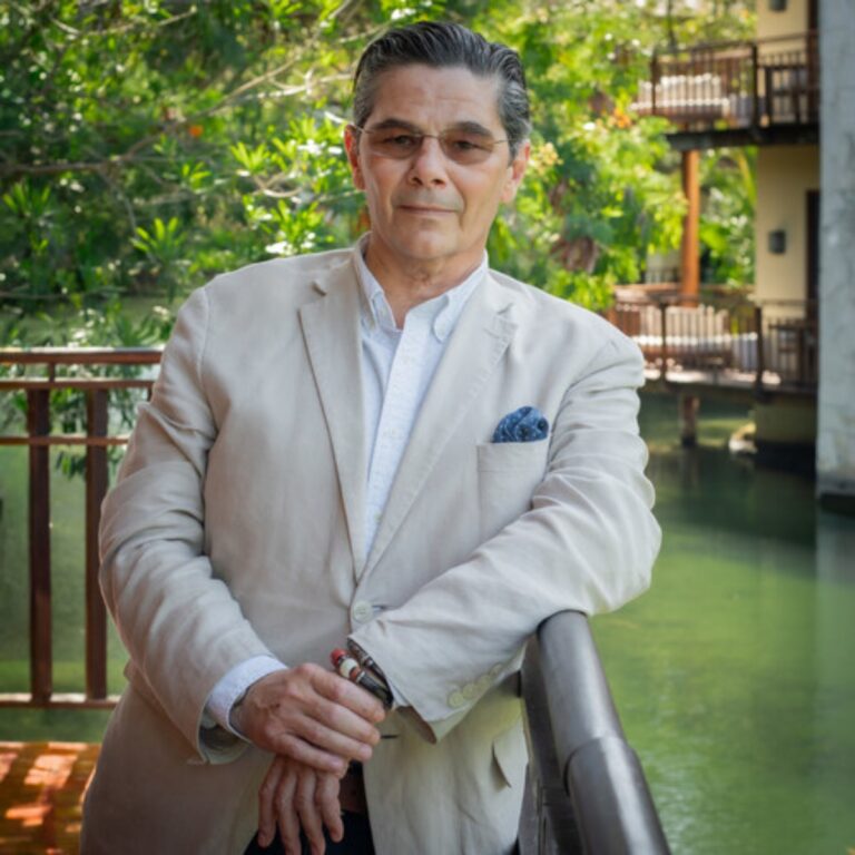 Man in a light beige blazer and white shirt leans on a railing beside a pond, with lush greenery in the background and a blue pocket square visible in his blazer pocket.