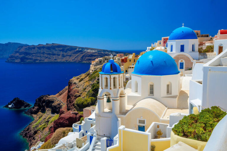 White buildings with blue domes perched on a cliff overlooking the deep blue Aegean Sea in Santorini, Greece.