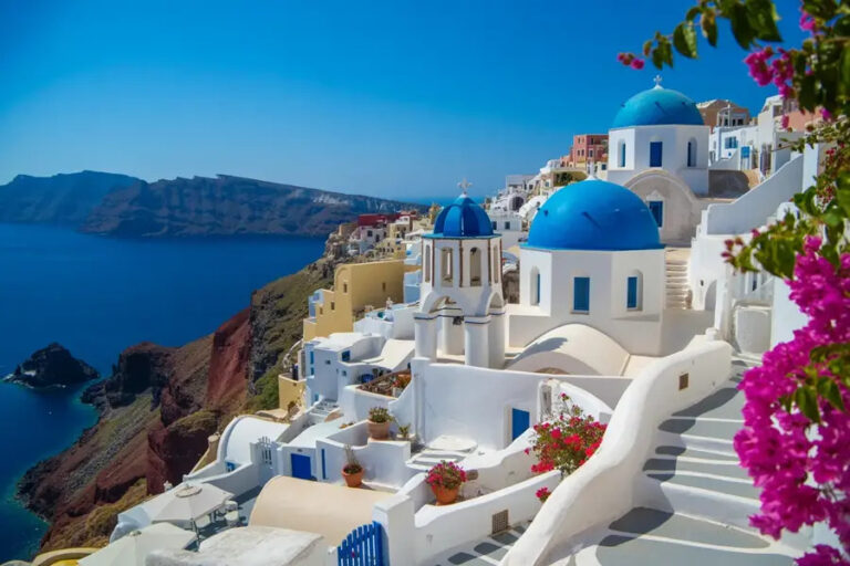 White-washed buildings with blue domes and stairs along a cliffside village overlooking the Aegean Sea in Santorini, Greece.