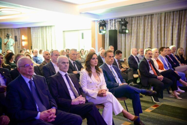 Audience seated in a conference hall, mostly professionals in business attire, listening to a presentation.