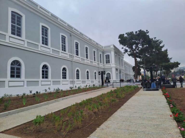 A pale gray, long historical building with arched windows, a central walkway, and people gathered near trees for an outdoor event.