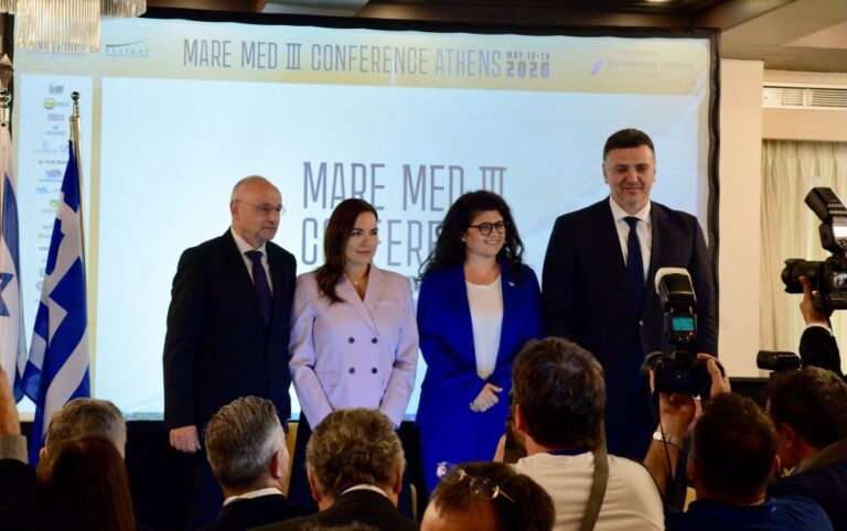 Four conference speakers pose for a photo on a stage with a projection screen and a Greek flag beside them.